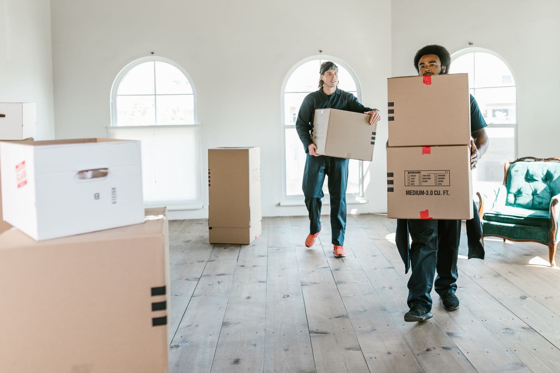 Zwei Umzugshelfer tragen gestapelte Kartons durch einen sonnigen Altbauraum mit Dielenboden